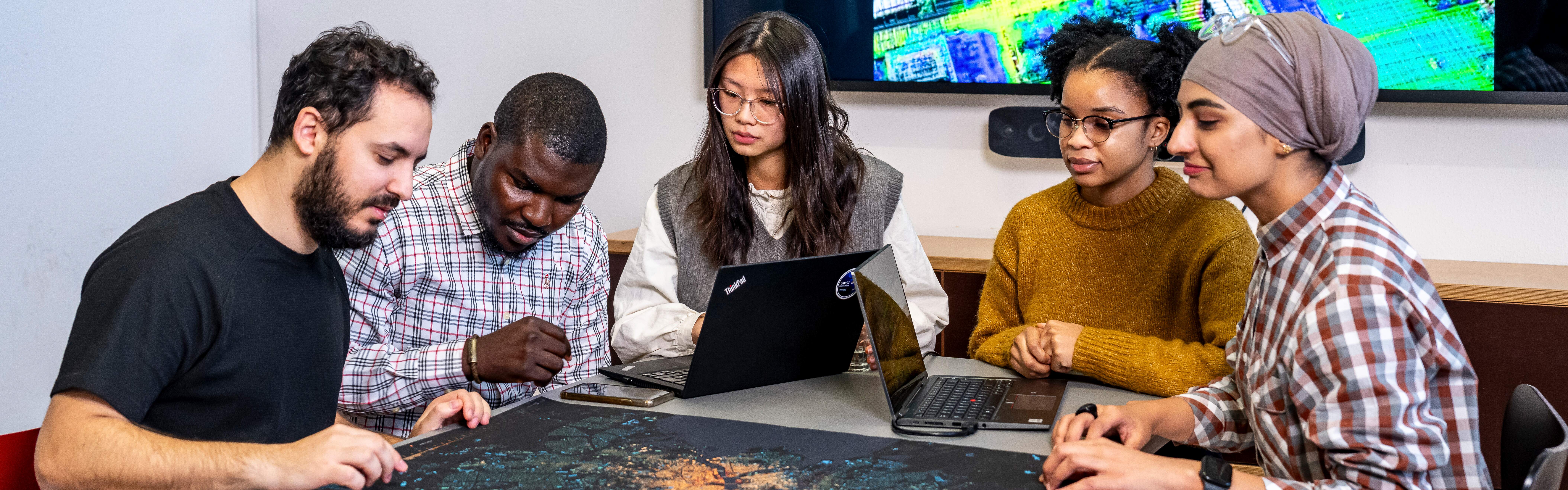 Several students sit together at a table and look at a map or a satellite image of a city from above. In the background is a screen on the wall showing a thermal image of a city from above.